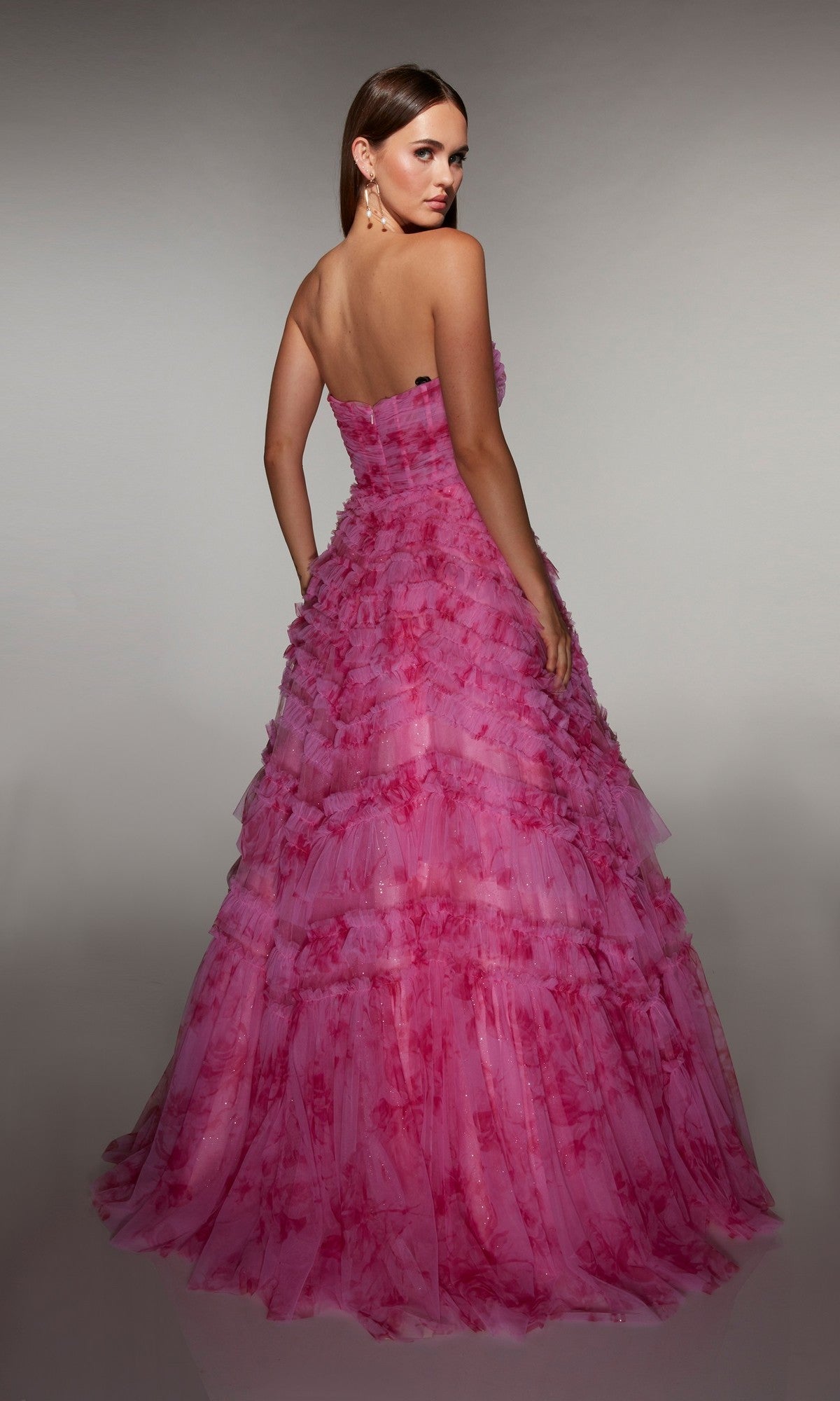 A woman models the Alyce 62164 Long Prom Dress—a strapless pink gown with a corset bodice, floor-length tiered ruffle skirt, and floral patterns—posing with her back slightly turned against a neutral background.